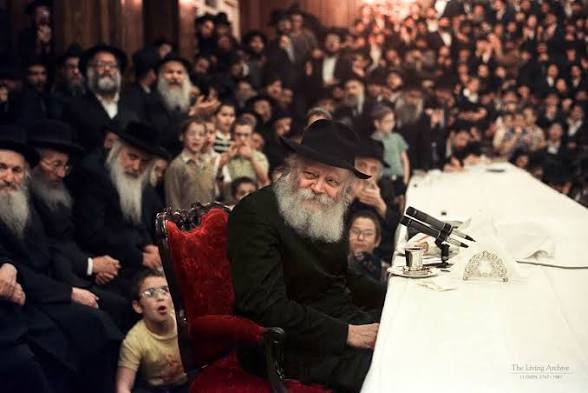The Lubavitcher Rebbe seated at a table at a farbrengen, with a large crowd of chassidim and children behind him
